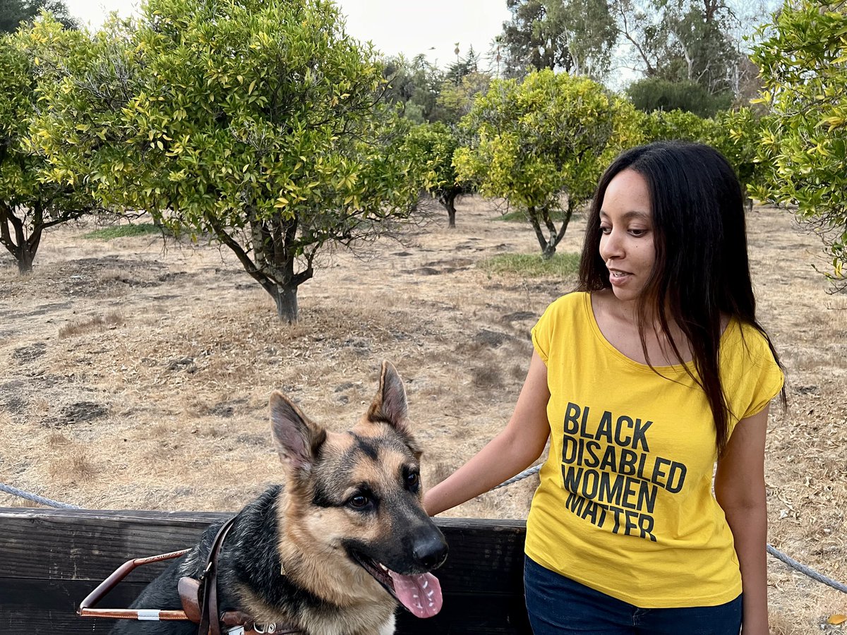 I’m a woman wearing a shirt that says Black Disabled Women Matter. My guide dog is sitting beside me and there are rows of trees behind us.