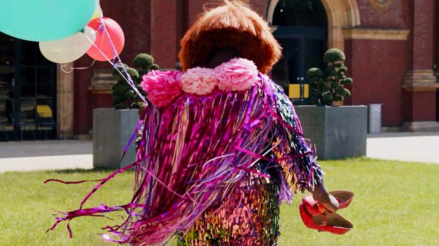 Person in a sharply cut orange wig, wearing a pink and purple sparkly cloak, holding balloons in one hand and sparkly red shoes in the other