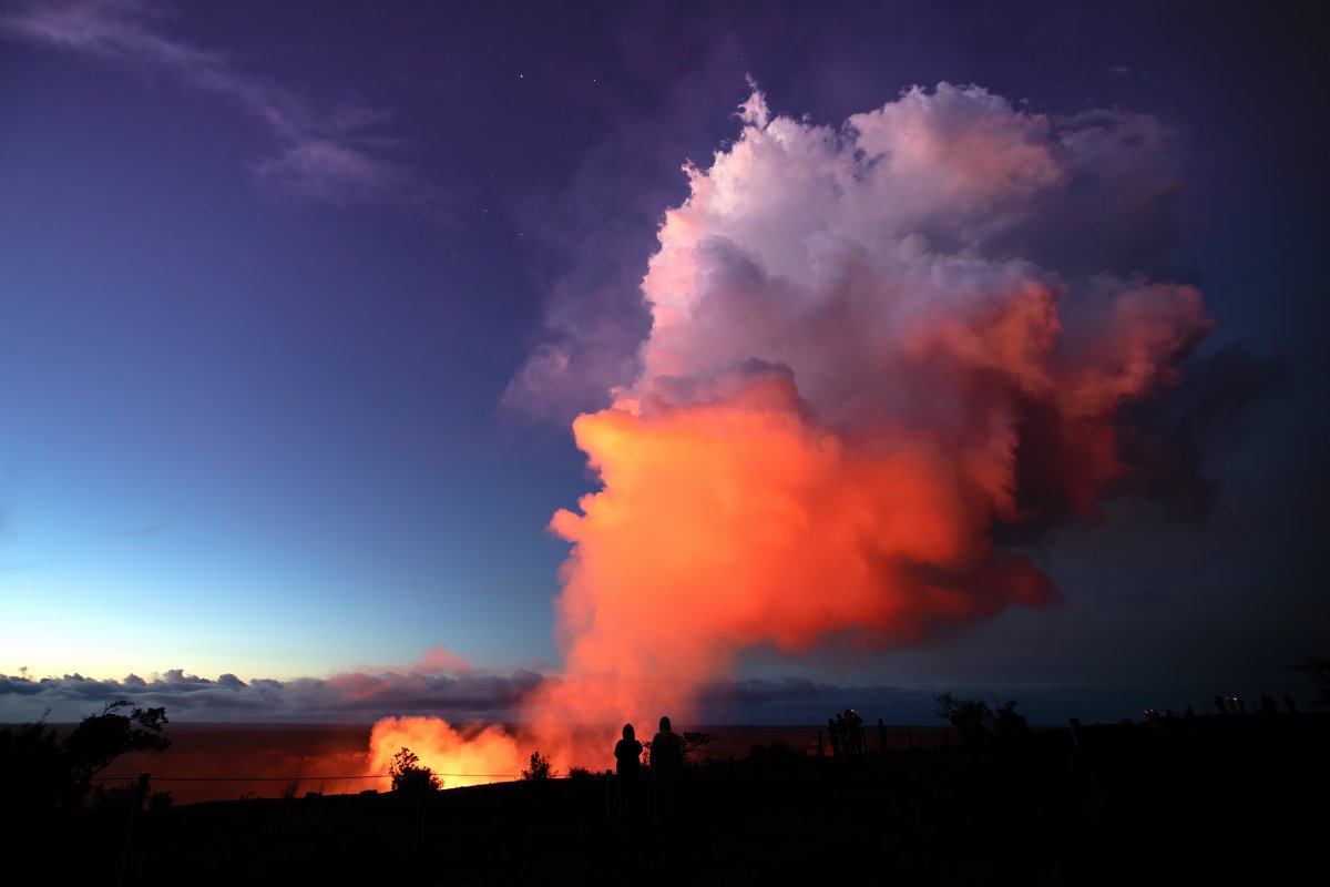 A large plume of gas billows out of a volcanic crater and is lit up with orange pinkish reflective glow from lava deep below. 
