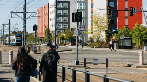 Two people dressed for cool weather walk along a sidewalk. Across the street are a number of condo buildings. A mass transit vehicle approaches the intersection. 