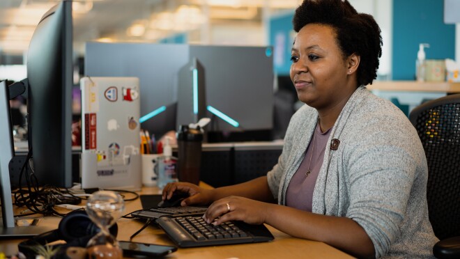 A woman sits at a computer, typing at her desk while looking at her keyboard. 