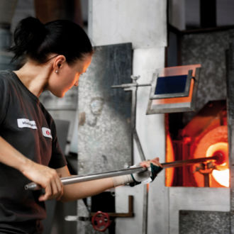 A woman holds a tool with a red-hot blob of glass at the end.