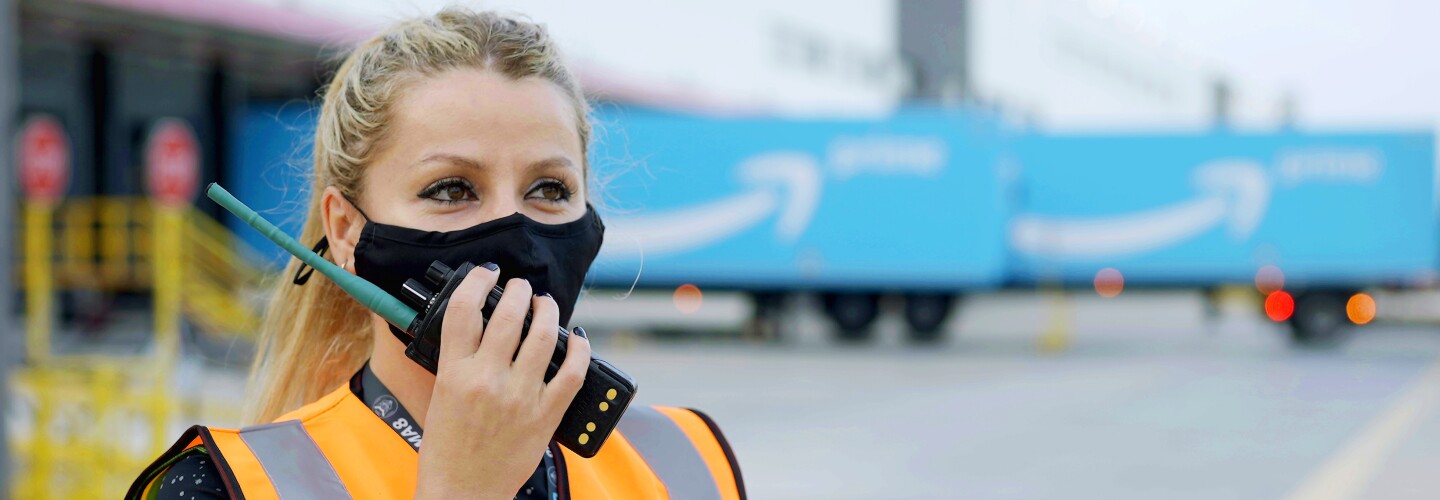 Ramona Moraru, supervisora de operaciones en la estación logística de Getafe. En el exterior del centro logístico con dos camiones de Amazon Prime de color azul con la sonrisa de Amazon en blanco. Ramona está hablando por un walkie talkie. Tiene el pelo claro, recogido en una cola, Con la mano derecha agarra el walkie talkie, tiene las uñas pintadas de negro. Lleva un tatuaje en la muñeca izquierda y un pulsera. Los ojos oscuros y maquillados y una mascarilla negra de Amazon. Lleva un chaleco de color naranja y un jersey negro con estrellas de color gris.  