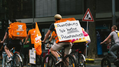 Demo von Lieferando-Fahrer:innen. Auf dem Demoschild steht: "Ausrüstung ist keine Privatsache"