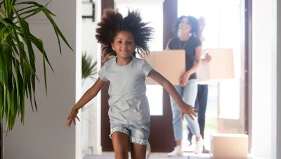 A little girl runs into a house.  Adults carrying moving boxes into the home are out of focus behind her.