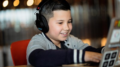 A boy wearing headphones works on a laptop at a coding camp while smiling.