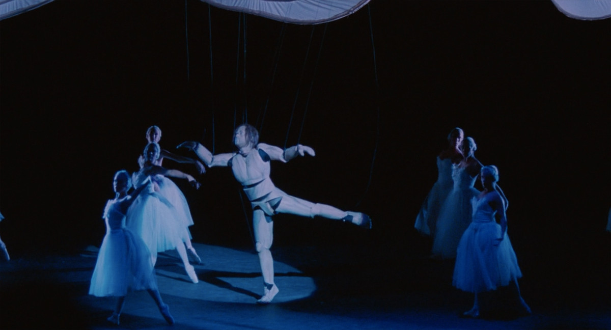 Still from the film "Being John Malkovich" picturing a life-sized marionette puppet dancing on a stage with ballerinas