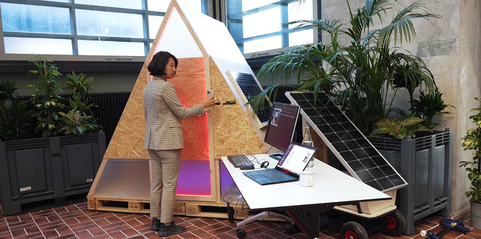 Eine Frau steht vor einem dreieckigen Gebäude mit Laptop und Solarpanel