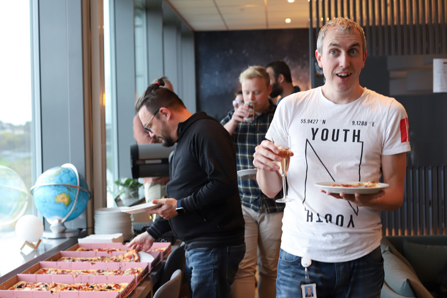 En smilende mann som holder champagneglass og tallerken med pizza mens andre står i kø bak