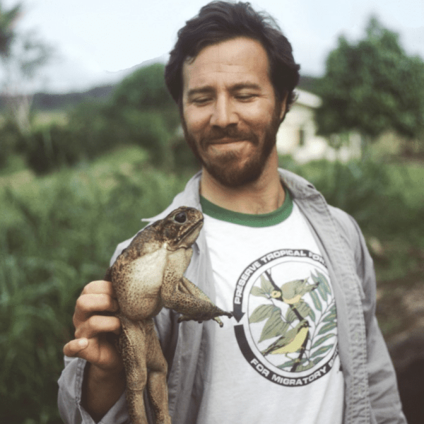 Mark Plotkin, smiling, holds up a toad, Bufo marinus, in his hand while standing in lush grass somewhere in Suriname.