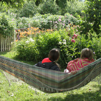 Two children sit in a hammock in a lush green garden.