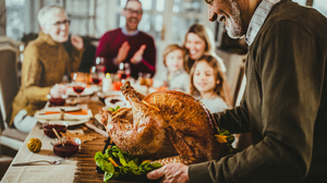 Older man carrying roast turkey to a dinner table. Focus is on man and turkey. Family around the dinner table blurred.