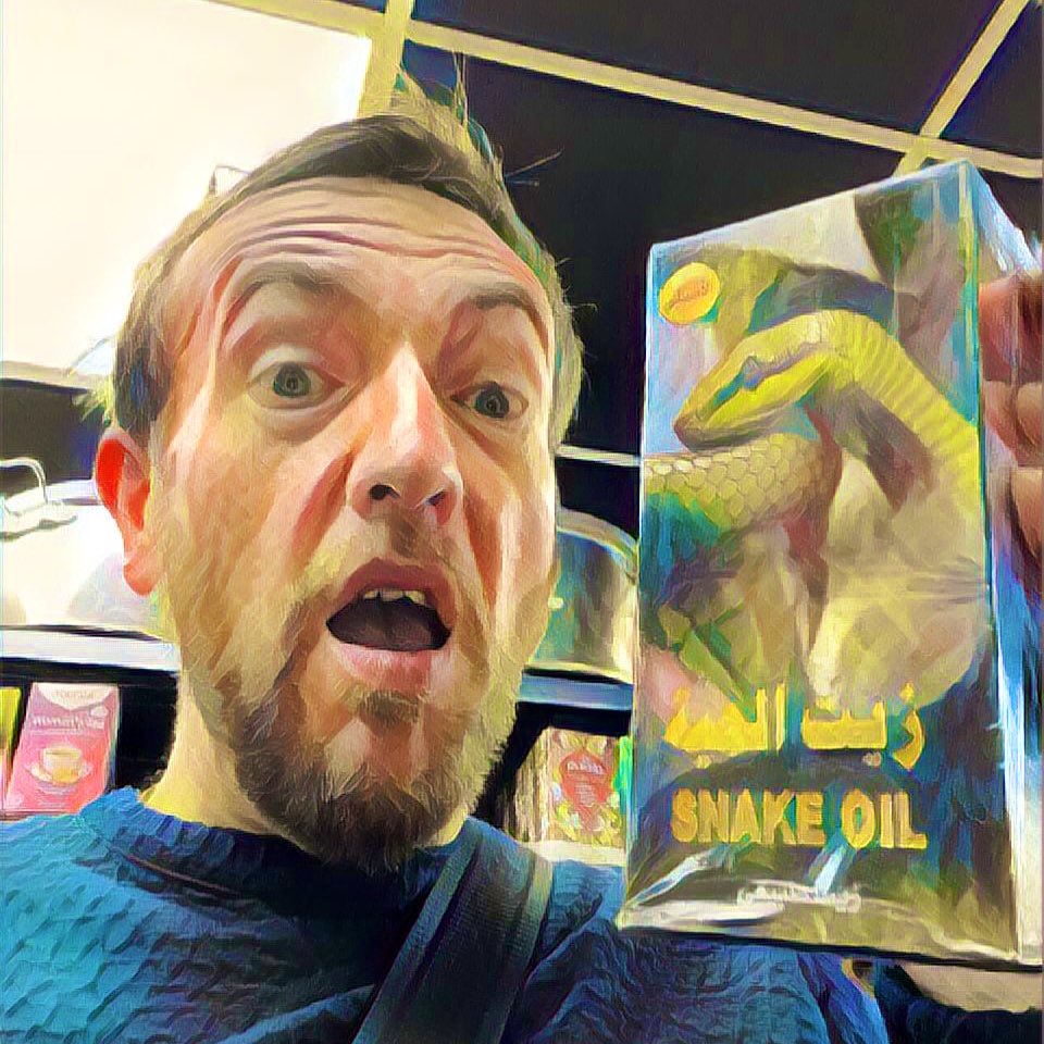 Neil a white male in his mid forties with sandy hair and a beard looks wide eyed and shocked at the box he is holding. The box has a green snake on the front and the words “Snake Oil”