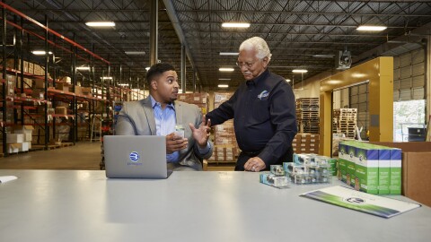 Two men speak to one another at a tabletop surface in a warehouse facility