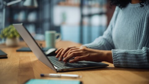 An image of a woman's hands typing on a laptop. She is sitting at a desk with a pen and paper and a plant and mug near her.