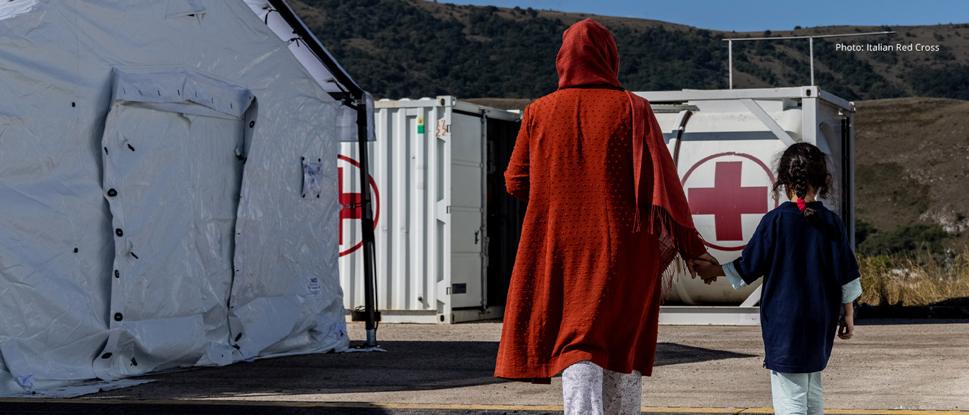 A woman holds the hand of a child, walking between emergency tents.