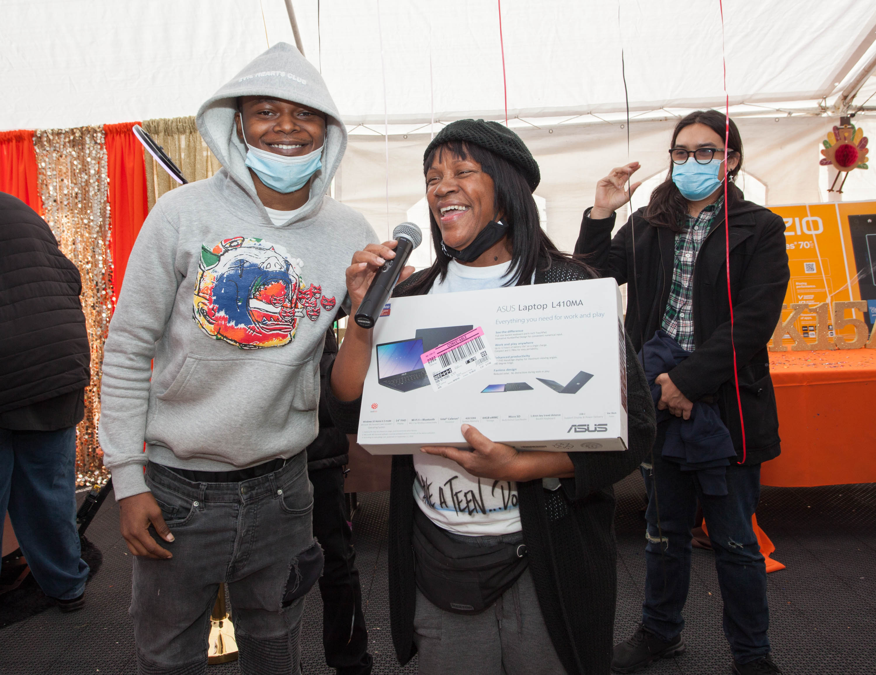 A woman smiling big holds a new laptop computer in her hands, appearing to be giving it to a young man to her right. The young man is wearing a grey hoodie, smiling wide in excitement.