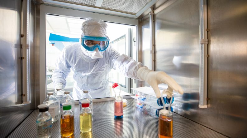 A scientist wearing a white protective suit and head covering reaches a gloved hand into an air-tight hood containing bottles of medicine