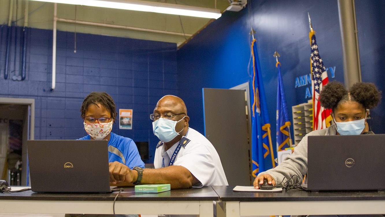 Two girls look at their computer screens while a man leans and points at one of the screens