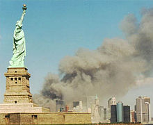 A monumental green copper statue of a woman with a torch stands on an island in front of a mainland where a massive plume of grey smoke billows among skyscrapers.