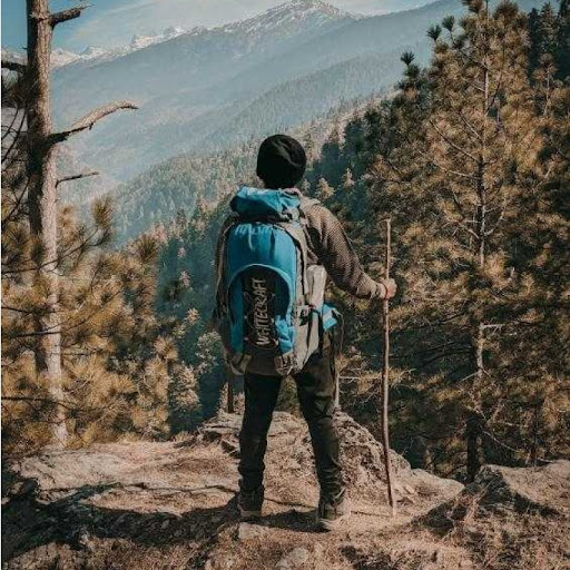 A young man wearing a turquoise backpack standing atop a vista overlooking the Himalayas.