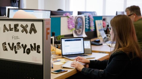 Two people sit at their desks which are side-by-side. There is a white board that reads "Alexa for everyone"