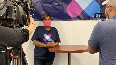 An image of a woman wearing a pink mask while smiling and leaning against a tall table. She is wearing a shirt showing she has been vaccinated and being filmed by a film crew. 