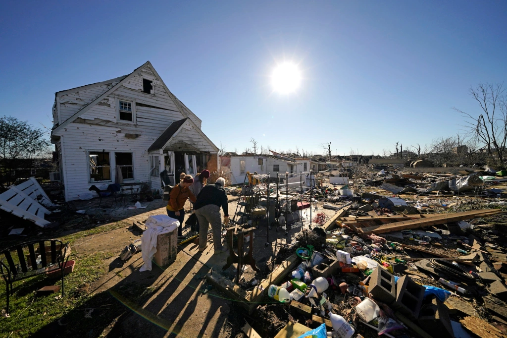 a white house with peeling paint and loose slats stands in front of a giant field of debris -- wood, bottles, and scrap metal -- through which people are walking. Overhead, a blazing sun looks down in a clear, blue sky.