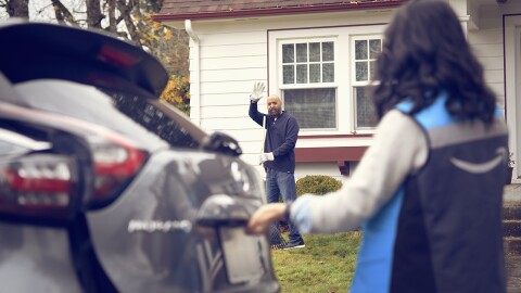 An image of a man waving from his yard. There is a woman wearing an Amazon delivery driver vest opening her trunk and looking back at him 