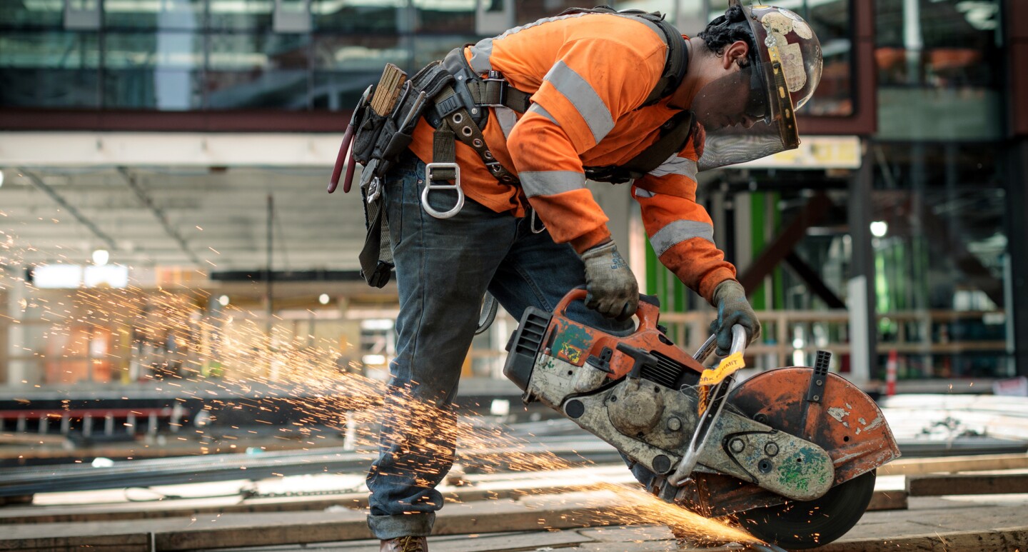 A man in a construction helmet and orange safety jacket is photographed at a construction site on Amazon's campus in Seattle, WA. He is using a circular saw to cut through metal. 