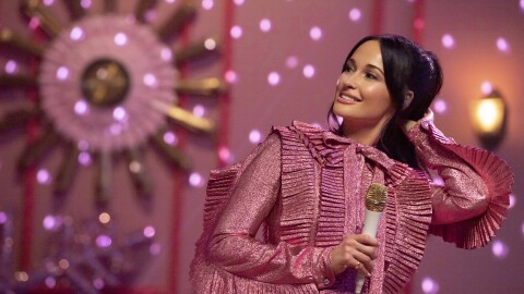 An image of Kacey Musgraves smiling while holding a microphone. She is dressed in a sparkly pink outfit and the room behind her is pink with twinkle lights lighting up the walls.