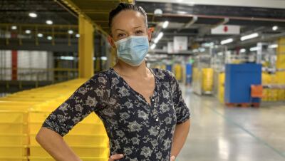 A woman wearing a mask stands in front of stacks of yellow plastic boxes. 