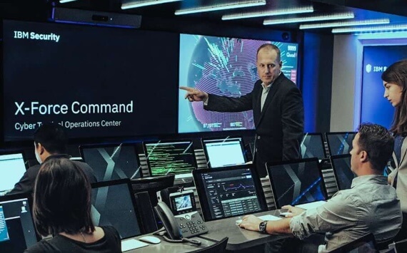 Four employees sitting in front of computers while a male executive is pointing at a large screen that reads IBM Security