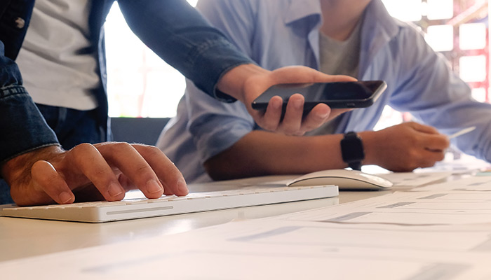 Person standing using keyboard and phone talking with seated person reviewing multiple documents