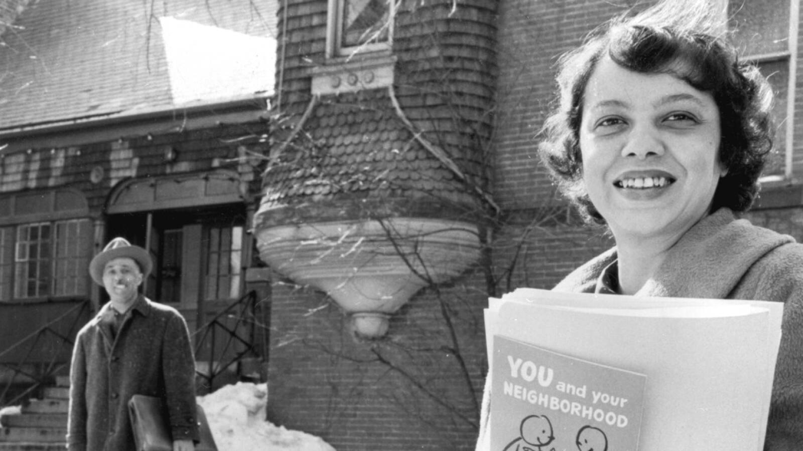 A black and white photo shows a woman in a winter coat holding a folder and book under one arm, and a man behind her in front of an old brick building.