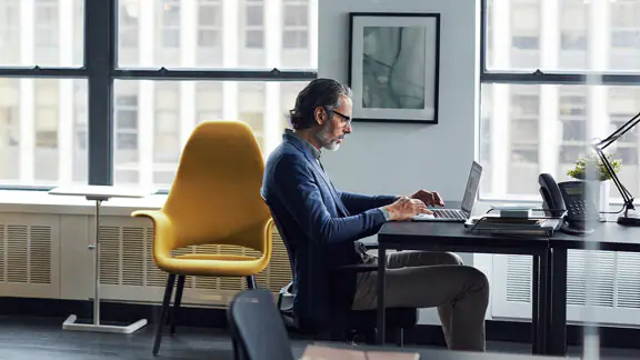 Person sitzt am Tisch und arbeitet am Laptop mit gelbem Stuhl im Hintergrund