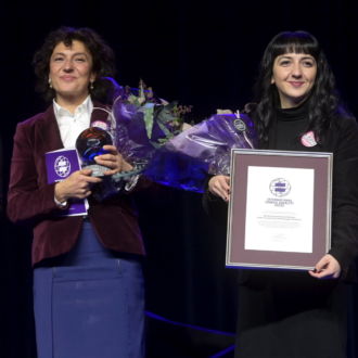 Three women stand next to each other, two of them holding flower bouquets and prizes.