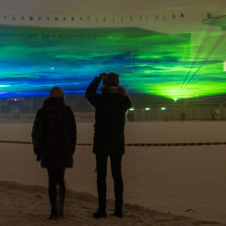 Two people are silhouetted in front of a light show that resembles the Northern Lights’ waves of purple, green and yellow colours.