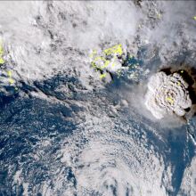 Satellite imagery of a Pacific Ocean location. Heavy, swirling white cloud cover hovers above the ocean, with a large, roughly circular formation of  dense cover on the left side of the image showing the volcano's eruption.