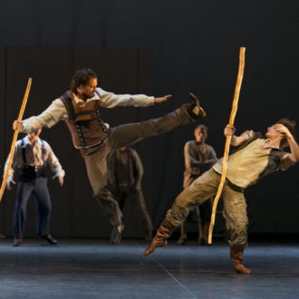 On a stage, one man jumps and kicks the air in front of another man.