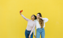 Two young women standing in front of a bright yellow wall taking a selfie together. 