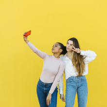 Two young women standing in front of a bright yellow wall taking a selfie together. 