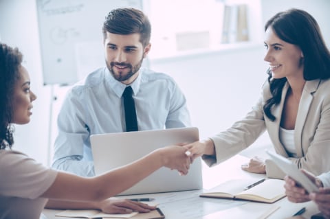 Two women shaking hands with smile while sitting at the office table with their coworker