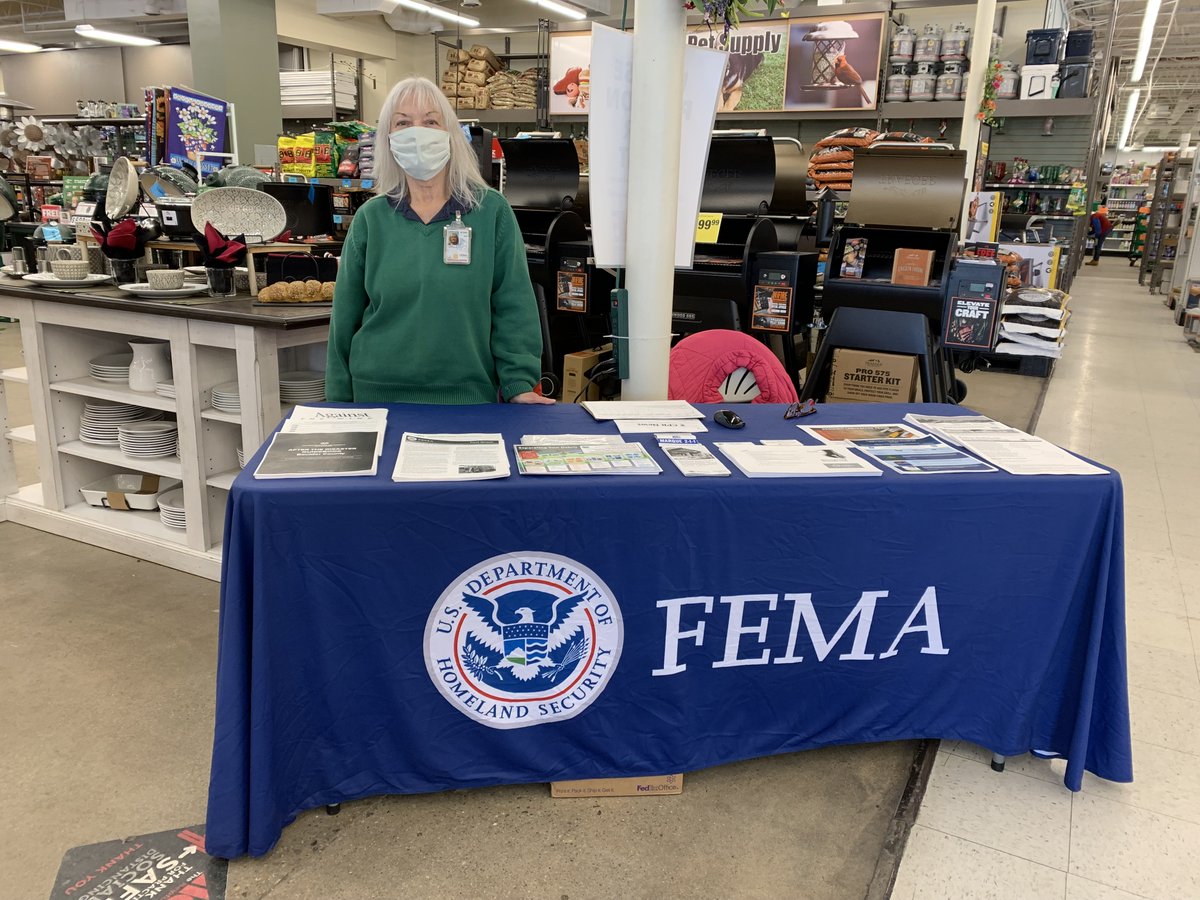 a photo of a woman standing in a green long sleeve sweatshirt behind a table with a blue tablecloth on it and the "FEMA" logo on the blue tablecloth