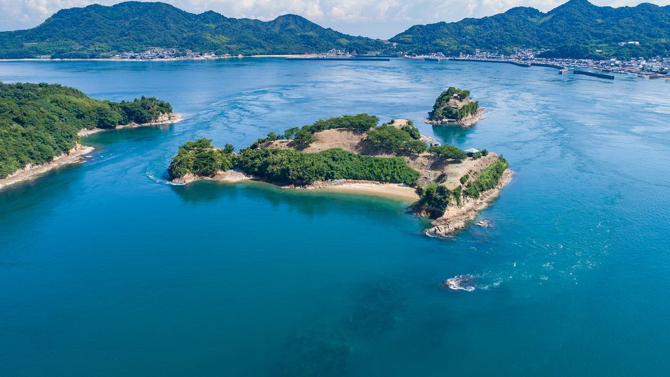 An aerial view of a small island, surrounded by water, with more land in the distance