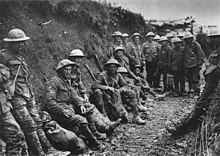 Black-and-white photo of two dozen men in military uniforms and metal helmets sitting or standing in a muddy trench.