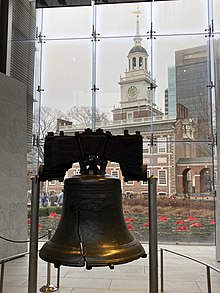 The Liberty Bell hangs in a glass-backed structure, with a brick, 18th-century building with a steeple visible in the background.