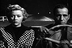 Black-and-white image of a man and woman, both with downcast expressions, sitting side by side in the front seat of a convertible. The man, on the right, grips the steering wheel. He wears a jacket and a pullover shirt. The woman wears a checkered outfit. Behind them, in the night, the road is empty, with a two widely separated lights way off in the distance.