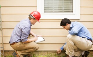 Repairmen, building inspectors, exterminators, engineers, insurance adjusters, or other blue collar workers examine a building/home's exterior wall and foundation. One wears a red hard hat and clear safety glasses and holds a clipboard. The other checks the foundation with tool.