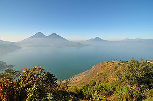 View across hills to a broad lake bathed in a light mist. The mountainous lake shore curves from the left foreground backwards and to the right, with several volcanoes rising from the far shore, framed by a clear blue sky above.